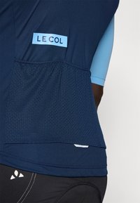 Maillot de cyclisme marine avec des accents bleus, poche en maille texturée et logo "LE COL" visible. Présente un design ajusté et un tissu respirant.