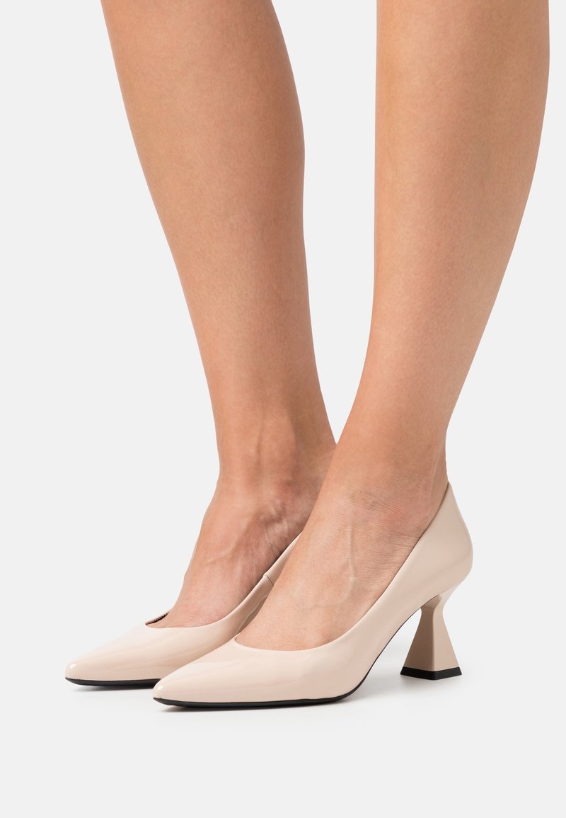 Escarpins beiges à bout pointu avec talons blocs évasés portés sur des jambes nues sur un fond uni.