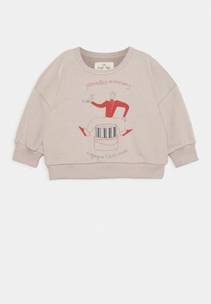 Hellbeiger Sweatshirt für Kleinkinder mit einer Grafik eines Mannes in roter Kleidung, der einen Vogel hält, sitzend auf gestreiften und einfarbigen Hüten, sowie französischem Text.