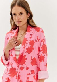 Květinový blejzr v jasně růžové barvě s výraznými červenými květinovými vzory. Má klopy s výstřihem, zapínání na jeden knoflík a vyhrnuté rukávy.