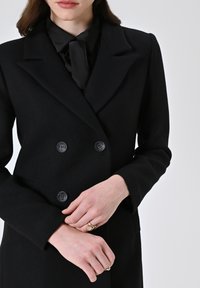 Cappotto doppiopetto nero realizzato in tessuto testurizzato, con grandi bottoni neri, revers a punta e vestibilità sartoriale. Indossato sopra una blusa nera.