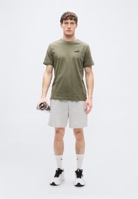 Λαδί βαμβακερό μπλουζάκι με κοντά μανίκια και μικρό λογότυπο, συνδυασμένο με γκρι σορτς, λευκές κάλτσες και μαύρα αθλητικά παπούτσια.