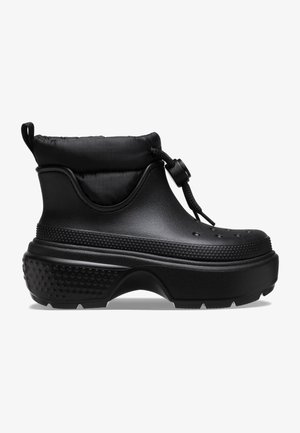 Botas negras hasta el tobillo con un puño acolchado, hechas de un material similar al caucho. Presentan acentos texturizados y una suela gruesa con dibujo para un mejor agarre.