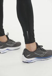 Grå sportskor med en meshinspirerad ovandel, vit mellansula och blå detaljer. Svarta leggings har en diskret reflexrand nära ankeln.