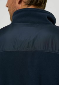 Giacca in pile blu navy con una patch in tessuto liscio e lucido sulla parte superiore della schiena. Presenta un colletto alto e cuciture piatte. Materiale testurizzato e caldo.