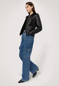 Černá péřová bunda s kapucí, prošívaná textura, v kombinaci s volně střiženými modrými džínovými cargo kalhotami se bočními kapsami a subtilními kovovými doplňky.