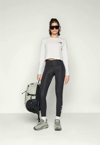 Fekete, magas derekú leggings, fehér, rövidített, hosszú ujjú felső, szürke hátizsák és szürke sportcipő; a modellen oversized fekete napszemüveg van.