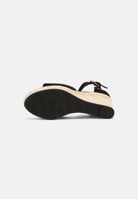 Sandale compensée noire avec semelle plateforme tissée beige et bride à la cheville, vue de dessous montrant la semelle extérieure en caoutchouc noir texturée.