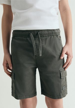 Persona con pantalones cortos cargo verde oscuro con cintura de cordón y bolsillos laterales, combinados con una camiseta blanca, de pie frente a un fondo liso.
