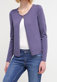 Cardigan tricoté violet avec col rond, fermeture éclair à l'avant, panneaux texturés et manches longues, associé à un haut blanc et un jean bleu.
