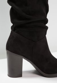 Bota de ante negra hasta la mitad de la pantorrilla con un tacón ancho y un caño ligeramente caído, mostrada de lado sobre un fondo claro.