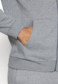 Main dans la poche avant d'un sweat à capuche gris zippé, assorti à un pantalon de survêtement gris.