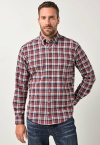 Man in een rood, blauw en wit geruit overhemd met lange mouwen en donkerblauwe spijkerbroek, staande tegen een effen achtergrond.