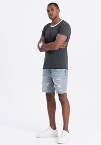Camicia grigia a maniche corte con colletto bianco, abbinata a pantaloni corti in denim chiari e strappati e scarpe da ginnastica bianche. Il modello è in piedi con le braccia incrociate.