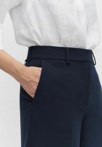 Pantalon bleu marine avec une texture lisse, présentant un devant plat et des poches latérales. Associé à une chemise blanche ample.