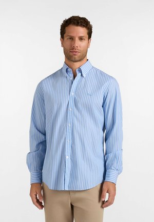Uomo che indossa una camicia a righe verticali azzurro chiaro e bianca con bottoni e pantaloni kaki, in piedi davanti a uno sfondo bianco uniforme.