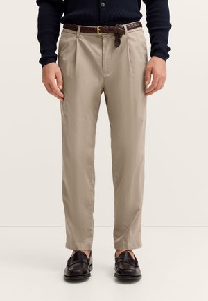 Mann trägt beigefarbene, plissierte Hose mit geflochtenem braunem Ledergürtel, schwarze Loafers und einen dunklen, gerippten Pullover, steht vor einem weißen Hintergrund.