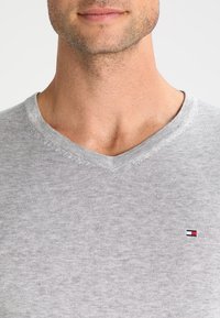 Pull en coton gris à col en V avec un tricot texturé. Présente un petit logo sur le côté gauche de la poitrine en rouge, blanc et bleu.