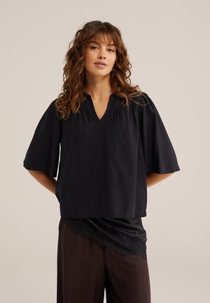 Žena s vlnitými hnedými vlasmi má na sebe voľnú čiernu blúzku s výstrihom do V a širokými rukávmi a tmavé nohavice, pózuje s rukami za chrbtom na neutrálnom pozadí.