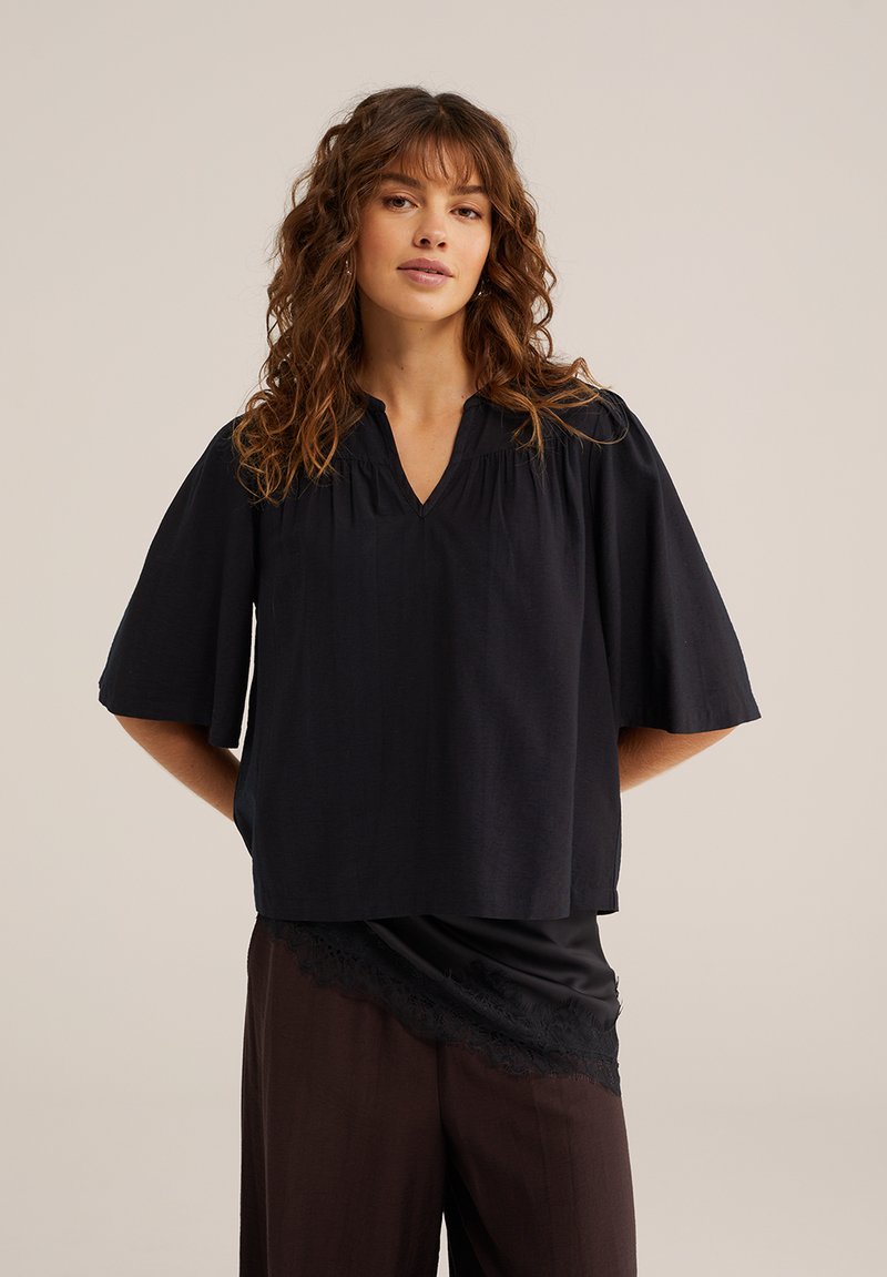 Barna hullámos hajú nő laza fekete V-nyakú blúzt visel széles ujjakkal és sötét nadrágot, semleges háttér előtt, kezeit a háta mögé téve pózol.