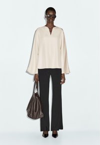 Blouse crème avec des manches larges et un décolleté en V, associée à un pantalon noir évasé. Le mannequin porte un sac marron foncé et des lunettes de soleil.