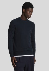 Marinblå stickad tröja med rund hals, långa ärmar och ribbad nederkant. Har en diskret logotyp på bröstet och en mjuk textur.