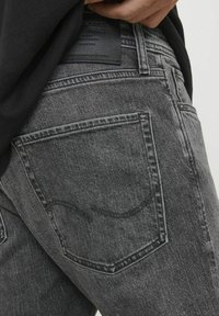 Vaqueros de mezclilla gris con una textura desgastada, que presentan dos bolsillos traseros y una etiqueta de cuero negro en la cintura, con detalles de costura visibles.