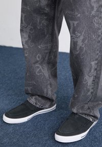 Sorte slip-on sko med hvit såle, teksturert, sammen med grå jeans med et subtilt mønster, plassert på en blå teppebakgrunn.