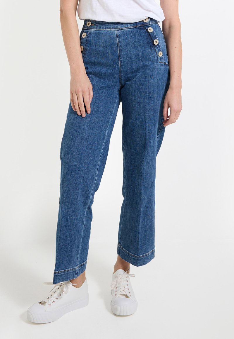 Pantalons en denim bleu taille haute avec une coupe droite, ornés de boutons sur les côtés et de ourlets retroussés, associés à des baskets blanches.