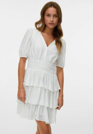 Robe blanche à volants avec un col en V et des manches courtes bouffantes. Elle présente une taille smockée et un tissu texturé, créant une silouhette fluide.