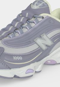Κοντινή λήψη ενός αθλητικού παπουτσιού New Balance 1000 σε ανοιχτό μωβ και άσπρο χρώμα, που δείχνει τα κορδόνια, το λογότυπο και τις υφασματικές λεπτομέρειες της σόλας.