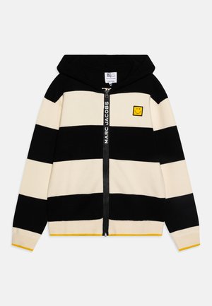 Cardigan rayé en noir et crème avec une fermeture éclair à l'avant, une capuche et un patch représentant un smiley jaune. Comprend des accents jaunes sur les poignets et l'ourlet.