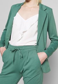 Zelený blazer s přiléhavým střihem, nošený přes bílou blůzu s volánky. Shodné kalhoty mají elastický pas a boční kapsy.