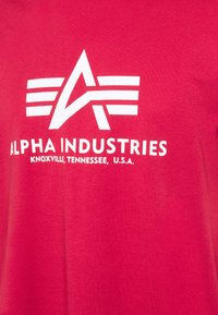 Rotes Baumwoll-T-Shirt mit einem großen weißen Logo, das stilisierte Buchstaben "A" und Streifen zeigt. Der Text lautet: "ALPHA INDUSTRIES, KNOXVILLE, TENNESSEE, U.S.A."