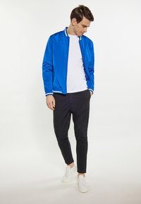 Jachetă de bombardier albastră cu detalii albe, purtată peste un tricou alb. Pantaloni negri skinny și adidași albi completează ținuta.