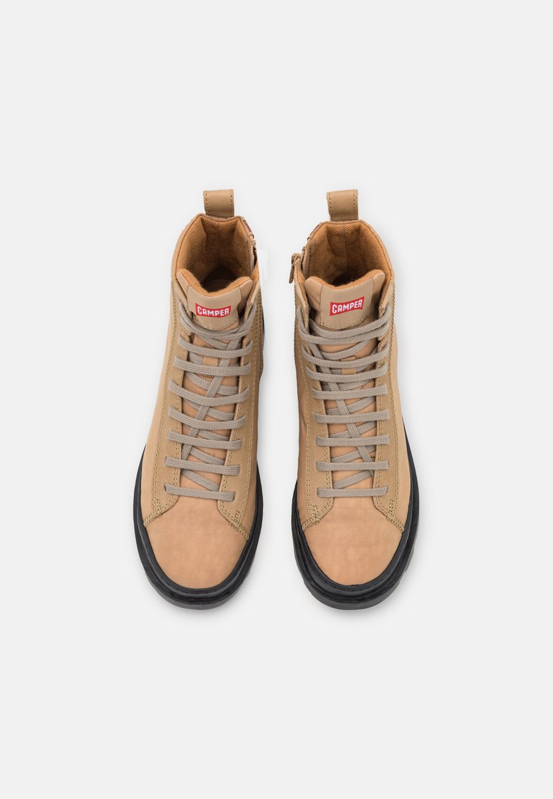 Beige Schnürsenkeln, schwarze Sohlen und roten Logo-Patches oben auf der Zunge, von oben betrachtet.