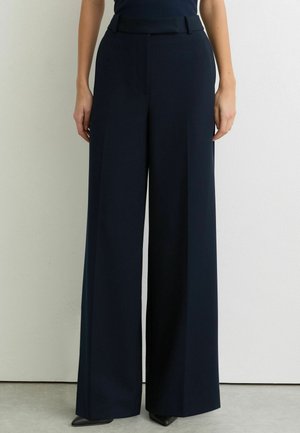 Mujer con pantalones azul marino de cintura alta y pierna ancha combinados con zapatos negros de punta afilada, de pie frente a una pared blanca lisa.