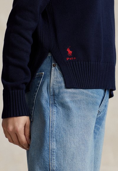 Tumši zils adīts džemperis ar ribotām aprocēm un apakšmalu, ar mazu sarkanu izšūtu logotipu. Valdāts kopā ar gaiši ziliem džinsu biksēm.