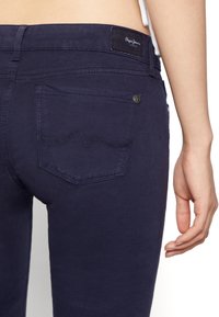 Jeans de mezclilla azul marino con una textura suave y un sutil detalle de costura en forma de onda en el bolsillo trasero. Presenta un parche de marca en la cinturilla.