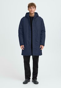 Temno modra dolga jakna s kapuco, z sprednjim zadrgo in gumbi na snap, nošena čez črn top in črne hlače. Gladka tekstura, sproščen dizajn.