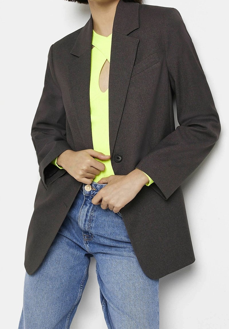 Tmavě šedý blejzr s klopovými límci, zapínáním na jeden knoflík a dlouhými rukávy, nošený přes jasně neonově zelený top a modré džínové kalhoty.