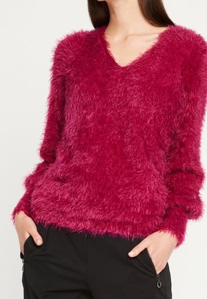 Femme portant un pull duveteux rose profond à col en V avec des manches longues et un pantalon noir avec des poches zippées, mains dans les poches.