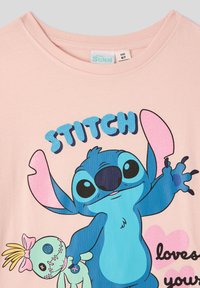 Jasnoróżowa bawełniana koszulka z niebieską postacią z kreskówki o dużych uszach, trzymającą zieloną lalkę. Napis brzmi "STITCH kocha twoje."