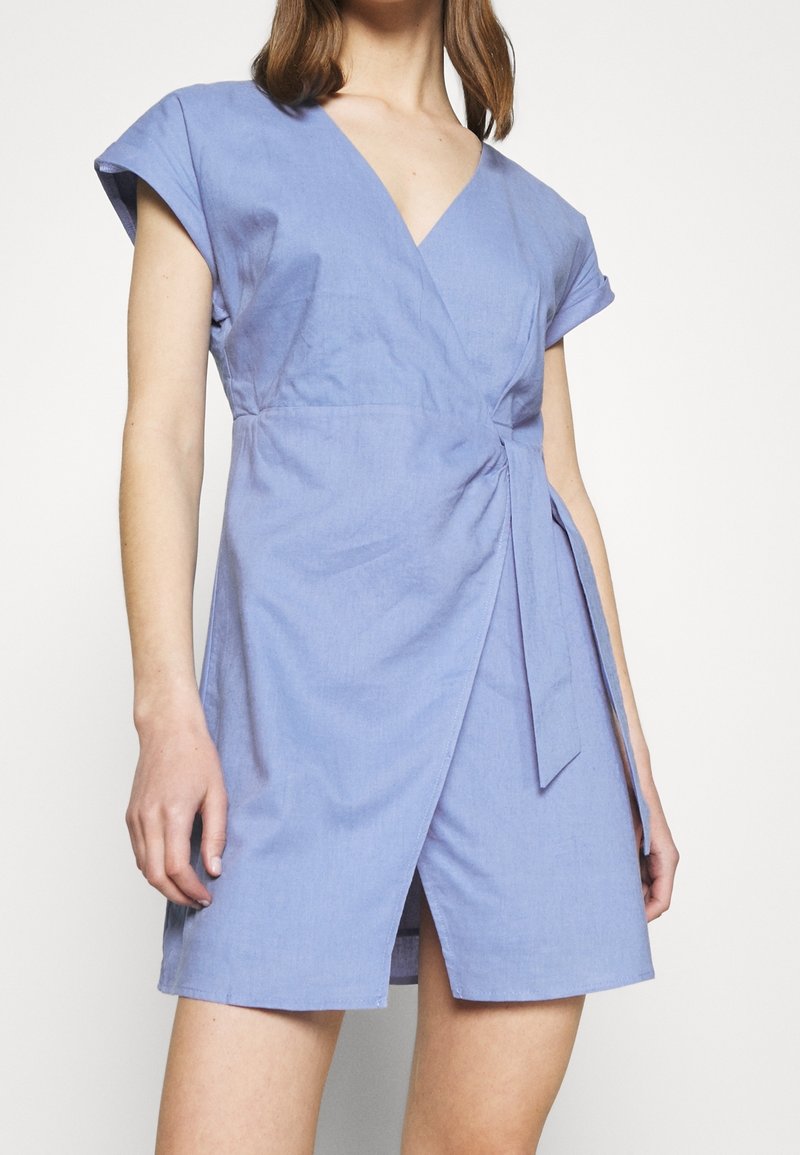 Femme portant une robe portefeuille bleu clair à manches courtes, avec un décolleté en V et un détail noué sur le côté, debout devant un fond blanc uni.