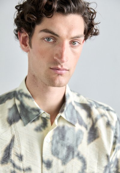 Cotton shirt with a light beige base, featuring a gray tie-dye pattern, short collar, and buttoned front, textured with subtle wrinkles.