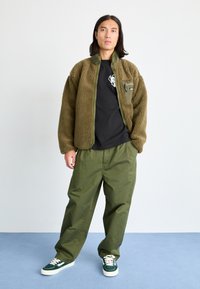 Puchowy oliwkowozielony kurtka z zamkiem błyskawicznym, przednia kieszeń; czarny t-shirt z grafiką; luźne oliwkowe spodnie; zielono-białe sneakersy.