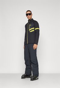 Veste zippée noire avec des accents jaunes et un logo, associée à un pantalon foncé et des bottes de ski. Modèle portant des lunettes de soleil, se tenant sur un fond uni.