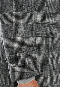 Szaro-czarna tkanina w kratę z teksturowanymi wzorami. Rękaw z mankietem ozdobiony czarnym guzikiem, kontrastującym z gładką podszewką.