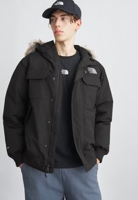 Jachetă neagră izolată, cu glugă căptușită cu blană, buzunare cu capse și manșete ribbate. Purtată peste un tricou negru și asortată cu pantaloni de culoare bleu deschis.