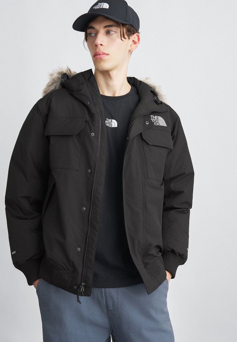 Jachetă neagră izolată, cu glugă căptușită cu blană, buzunare cu capse și manșete ribbate. Purtată peste un tricou negru și asortată cu pantaloni de culoare bleu deschis.